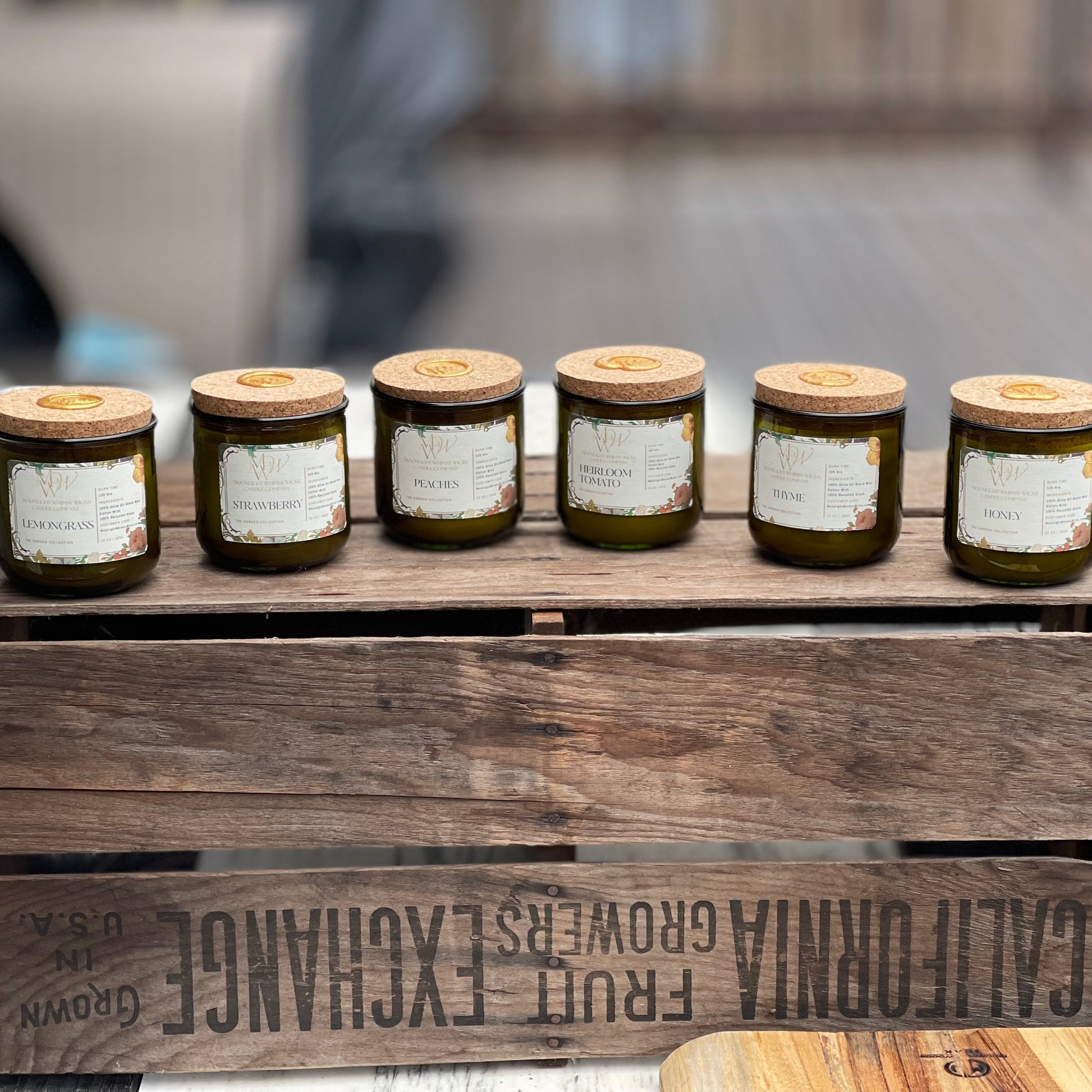Full lineup of Moonlight Whimsy Wicks’ Garden Collection candles displayed on a rustic wooden crate. Features eco-luxury scents: Lemongrass, Strawberry, Peaches, Heirloom Tomato, Thyme, and Honey. Each candle is hand-poured in a recycled green glass vessel with a cork lid and gold wax seal—perfect for garden lovers and sustainable home fragrance seekers.