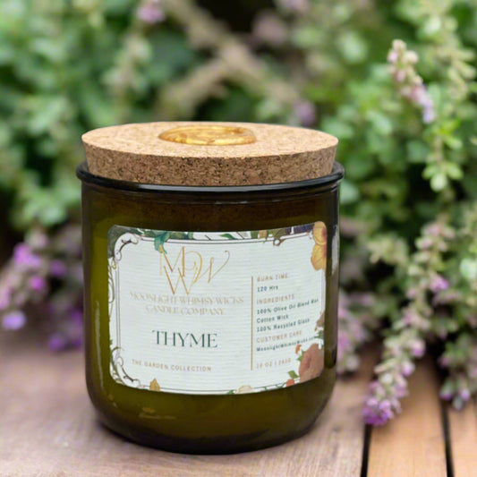 Thyme Candle from Moonlight Whimsy Wicks’ Garden Collection, displayed in a green recycled glass jar with a cork lid and floral label. Set against blooming thyme with purple blossoms, this eco-friendly olive oil wax candle evokes a fresh, herbal garden fragrance.
