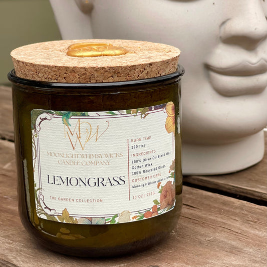 Lemongrass Candle by Moonlight Whimsy Wicks, featuring an olive oil wax blend in a green recycled glass jar with a cork lid and floral label. Set on a rustic wooden table beside a serene stone sculpture, this eco-friendly candle is part of the Garden Collection.