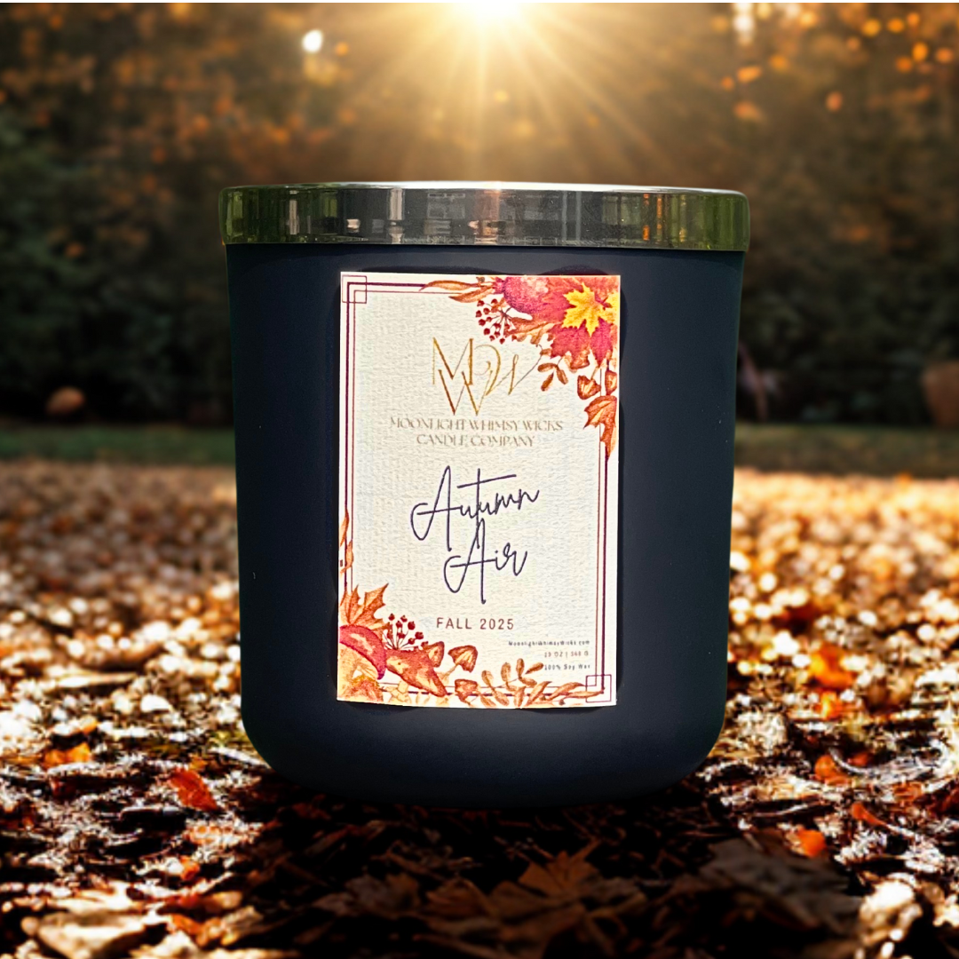 Autumn Air soy candle by Moonlight Whimsy Wicks, in a matte black jar with fall floral label, sitting on leaf-covered ground with golden sunlight beaming through the trees.