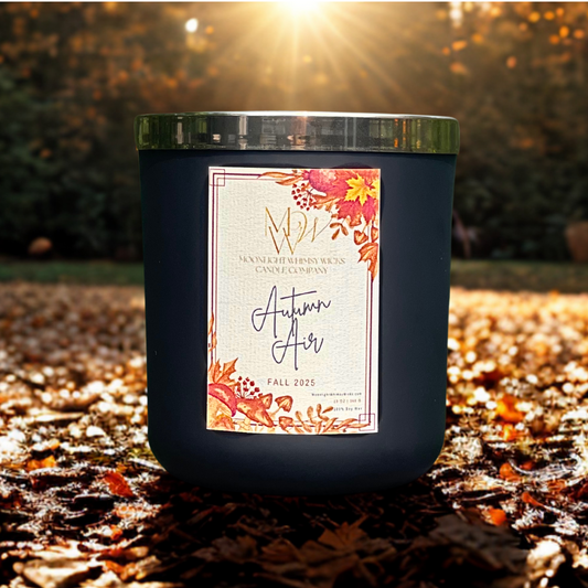 Autumn Air soy candle by Moonlight Whimsy Wicks, in a matte black jar with fall floral label, sitting on leaf-covered ground with golden sunlight beaming through the trees.