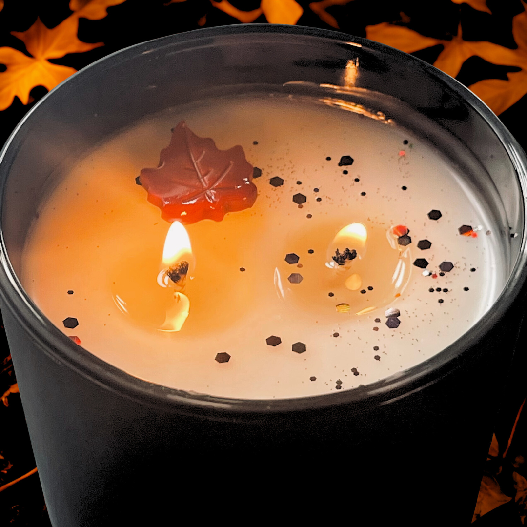 Close-up of a lit soy wax candle in a black jar, featuring two cotton wicks, fall-colored glitter, and a red maple leaf wax embed, creating a cozy autumn ambiance.