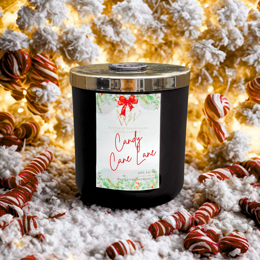 Luxury peppermint soy candle in matte black jar with gold lid, labeled “Candy Cane Lane” by Moonlight Whimsy Wicks, surrounded by red and white candy canes and snow-dusted holiday decor. Festive winter scene with warm golden Christmas lights.