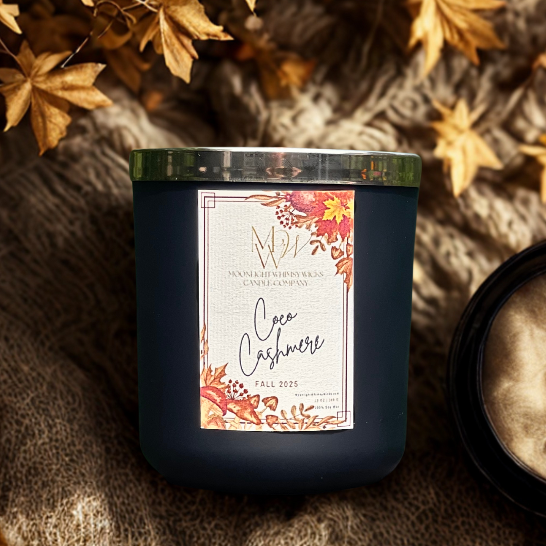 Coco Cashmere soy candle by Moonlight Whimsy Wicks, in a matte black jar with a fall floral label, styled atop a soft knit blanket with golden leaves and warm lighting.