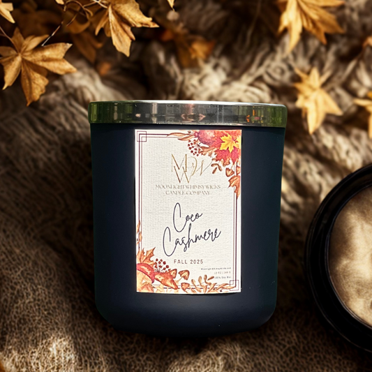 Coco Cashmere soy candle by Moonlight Whimsy Wicks, in a matte black jar with a fall floral label, styled atop a soft knit blanket with golden leaves and warm lighting.