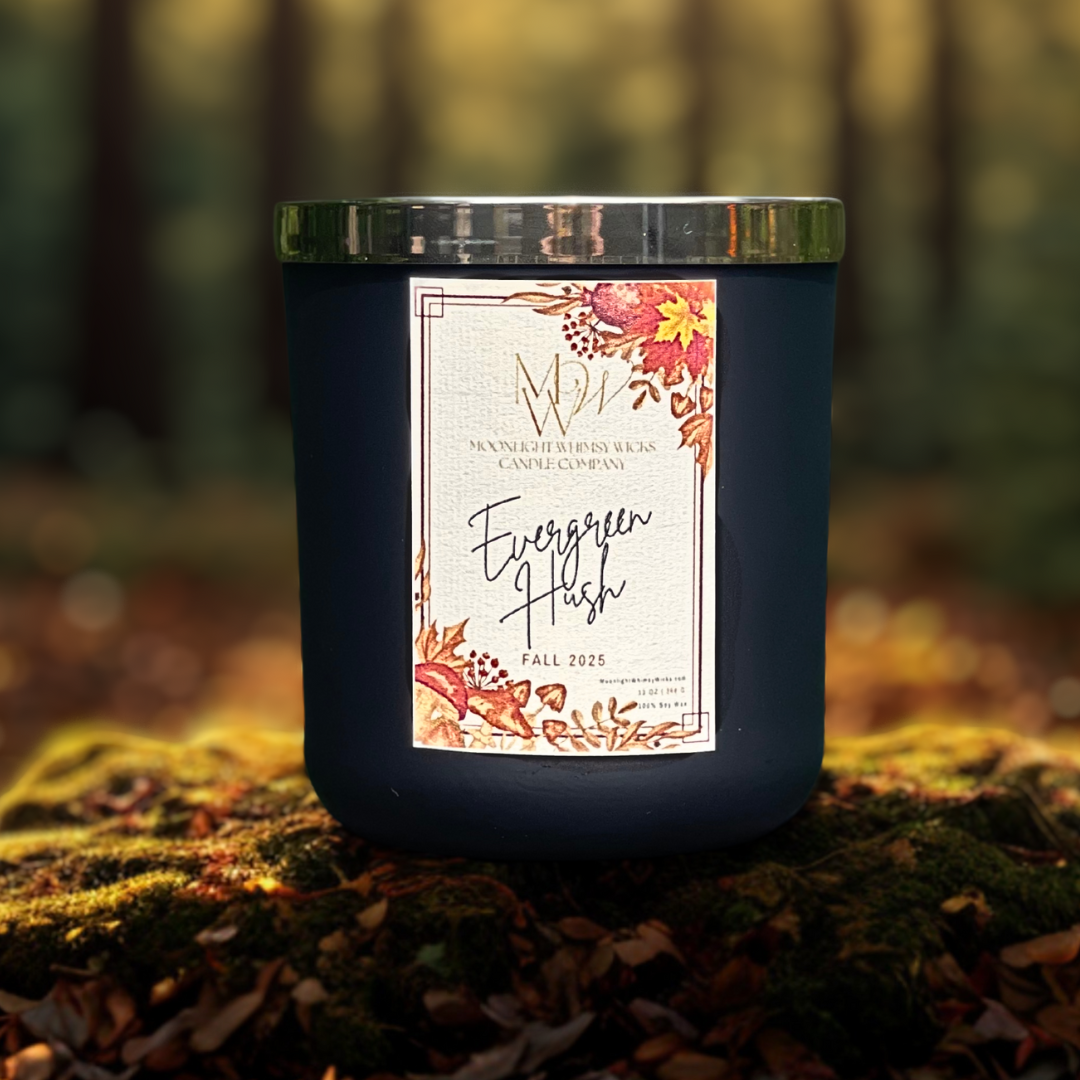 Evergreen Hush candle by Moonlight Whimsy Wicks, shown in a matte black jar with fall floral label, photographed outdoors on a mossy forest floor with blurred autumn trees in the background.