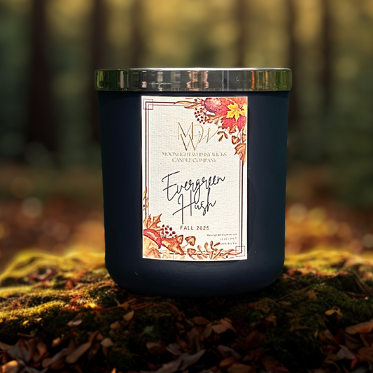 Evergreen Hush candle by Moonlight Whimsy Wicks, shown in a matte black jar with fall floral label, photographed outdoors on a mossy forest floor with blurred autumn trees in the background.