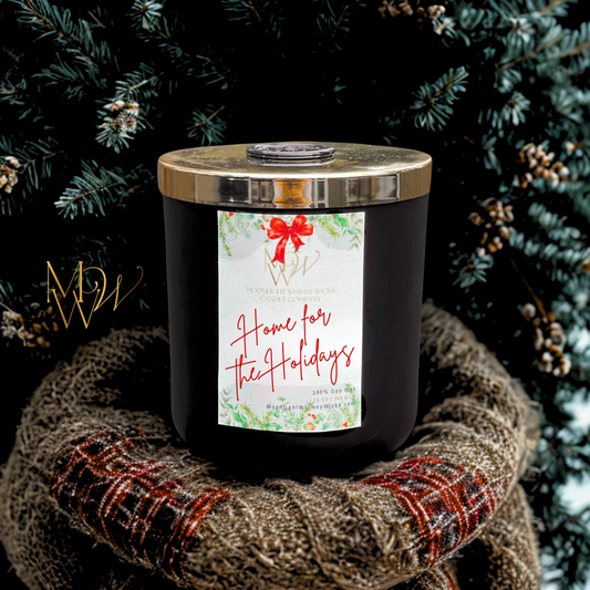Home for the Holidays candle by Moonlight Whimsy Wicks in matte black jar with gold lid and wax seal, nestled on a plaid wool scarf in front of a frosted evergreen tree. Handcrafted 100% soy wax Christmas candle featuring bayberry, clove, and orange zest—perfect for cozy winter décor, nostalgic gifts, and holiday home rituals.