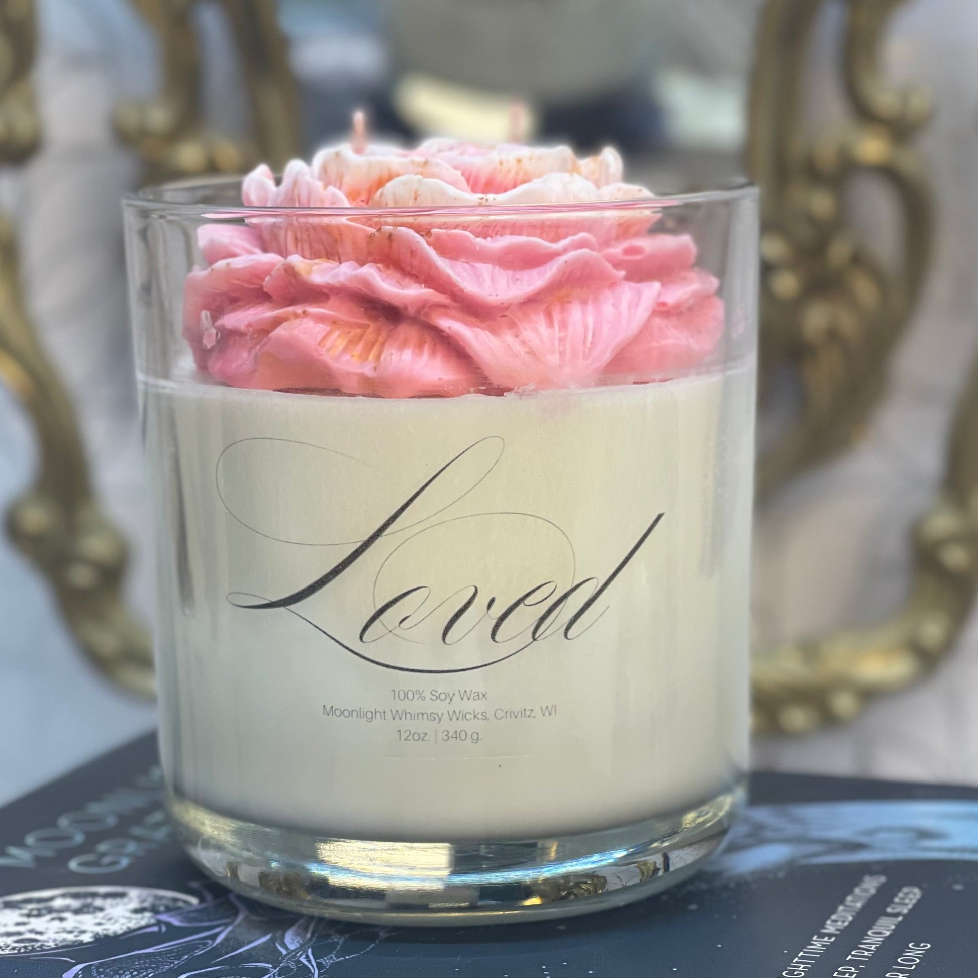 Candle with pink flowers in a glass holder on top of books, with a decorative chair and floral arrangement in the background.