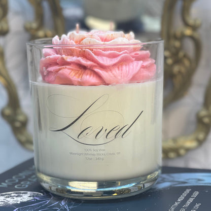 Candle with pink flowers in a glass holder on top of books, with a decorative chair and floral arrangement in the background.