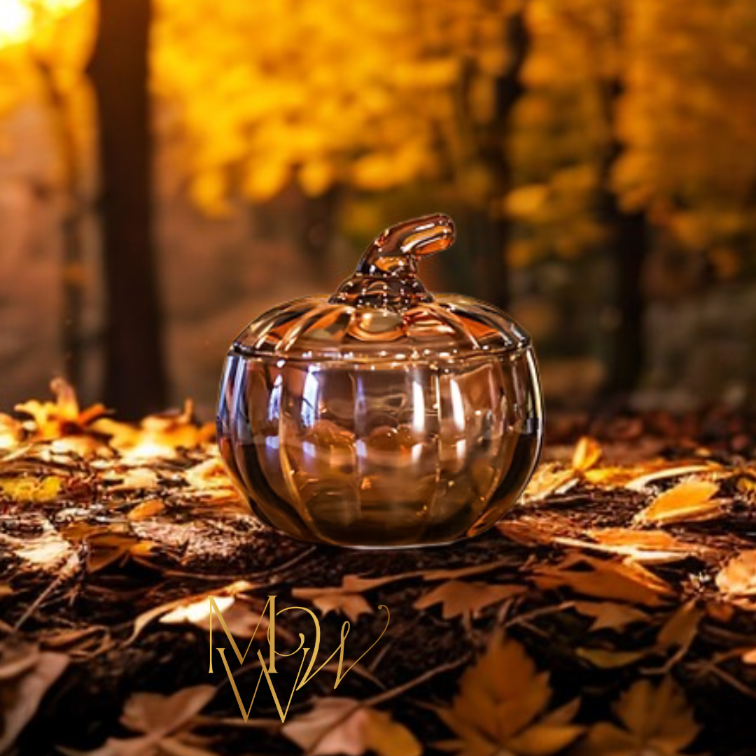 Glass pumpkin Candle with a lid on a leaf-covered ground with blurred autumn trees in the background