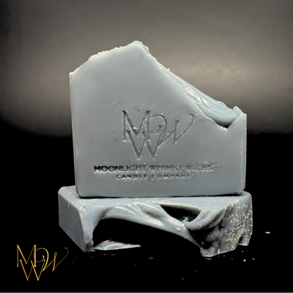 A bar of handmade soap in a blue gray color with a broken, rustic edge, featuring an embossed logo and the text 'MOONLIGHT WHIMSY WICKS Candle Company'. The soap is displayed on a black surface with a dark gray gradient background.