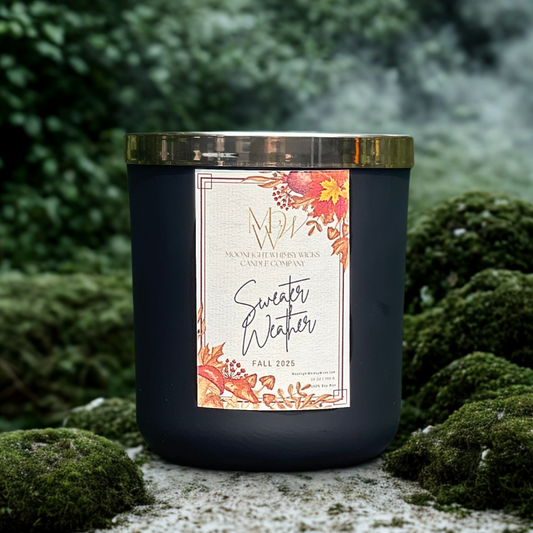 Sweater Weather candle by Moonlight Whimsy Wicks in a matte black jar with autumn floral label, styled on moss-covered stone with a misty forest background.