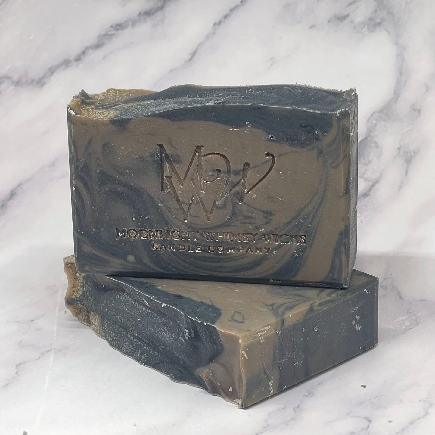 Two bars of brown and black Moonlight Whimsy Wicks soap with a logo on a marble surface