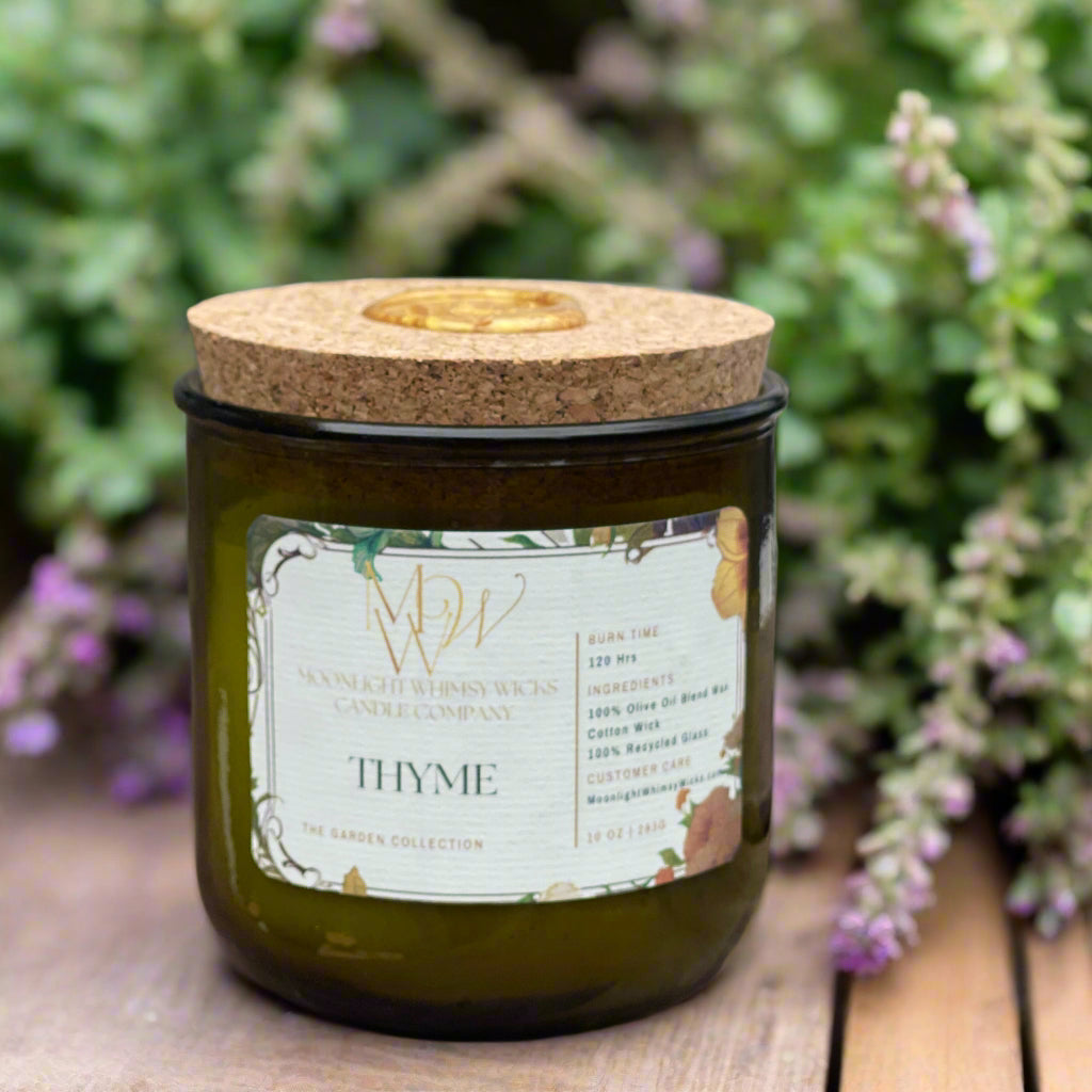 Thyme Candle from Moonlight Whimsy Wicks’ Garden Collection, displayed in a green recycled glass jar with a cork lid and floral label. Set against blooming thyme with purple blossoms, this eco-friendly olive oil wax candle evokes a fresh, herbal garden fragrance.