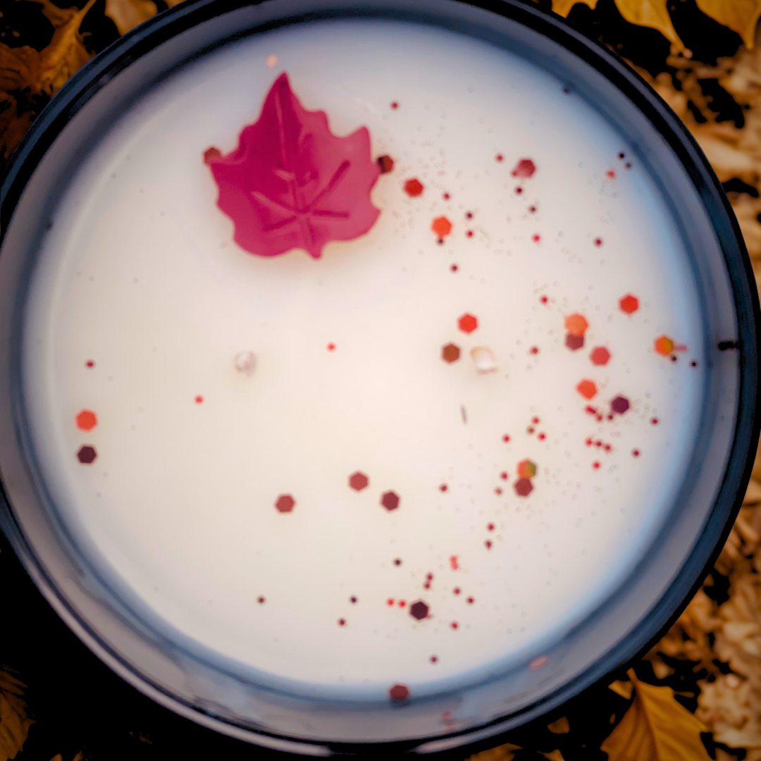 Top-down view of an unlit fall soy candle with red and orange glitter accents and a decorative red maple wax embed, in a sleek black vessel.