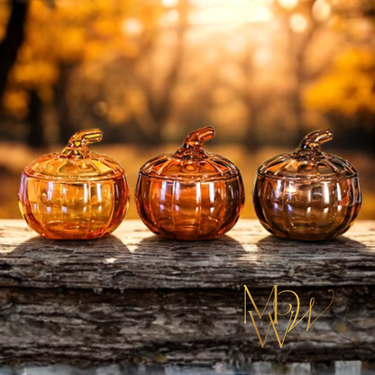 Three glass pumpkin candle containers on a wooden surface with a blurred autumn background.