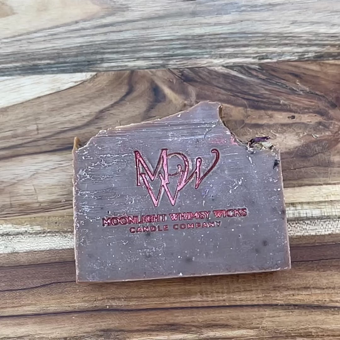 Short, close up video of Handcrafted rose clay artisan soap bar from Moonlight Whimsy Wicks, stamped with a red foil logo and topped with dried botanicals. Cold processed, natural soap made in small batches with moisturizing oils and gentle clays for radiant, sensitive skin.  Fragrance is inspired by BBW Dark Kiss